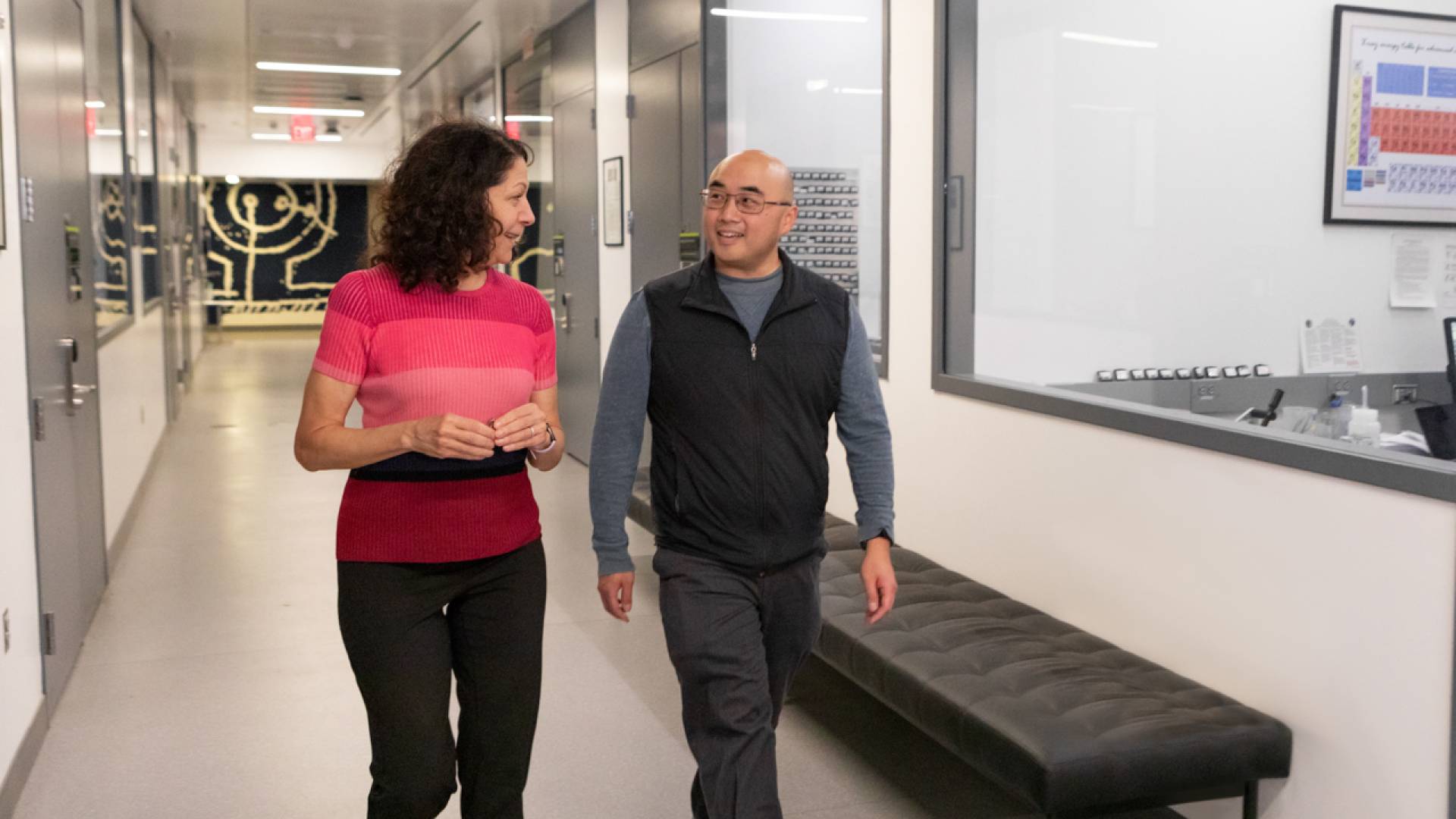 A man and woman walk past picture windows lining a long hall.
