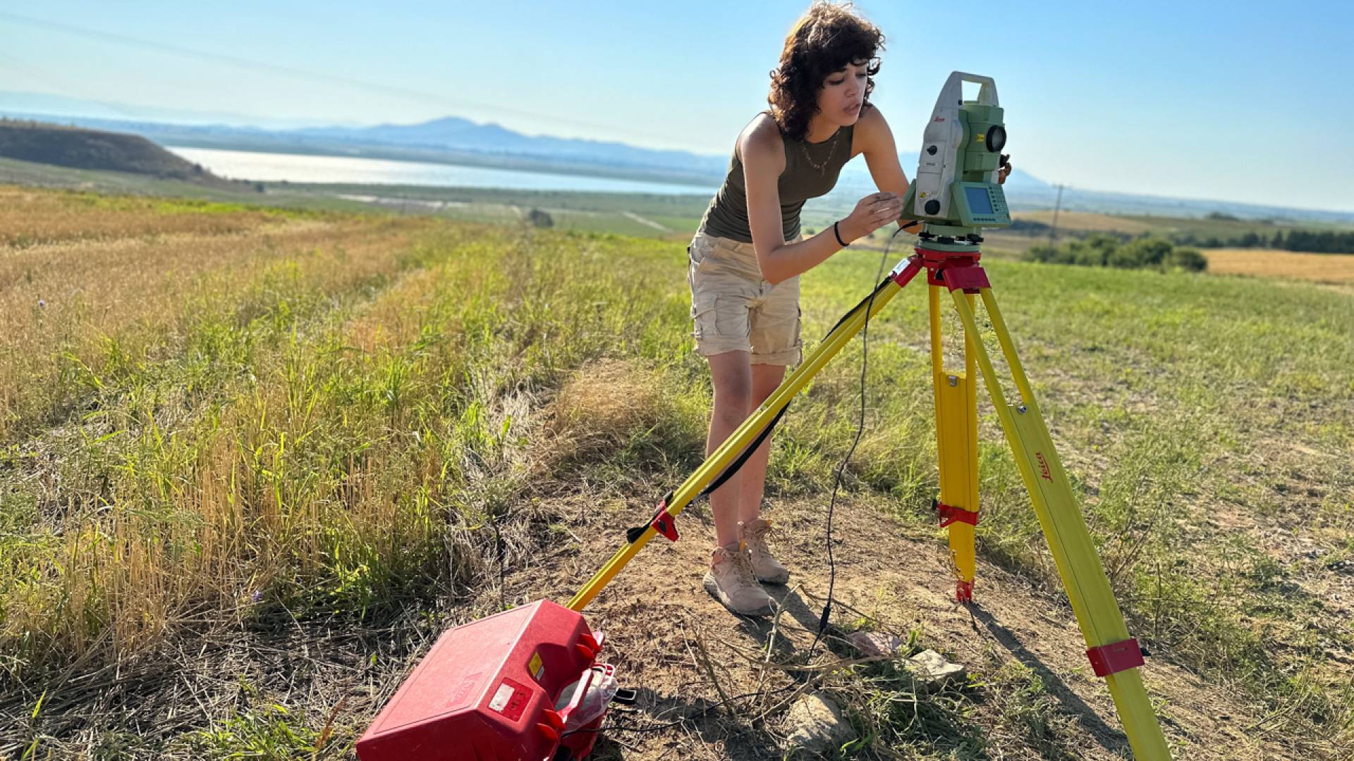 A students using an instrument to survey the land in the middle of a field of grass.