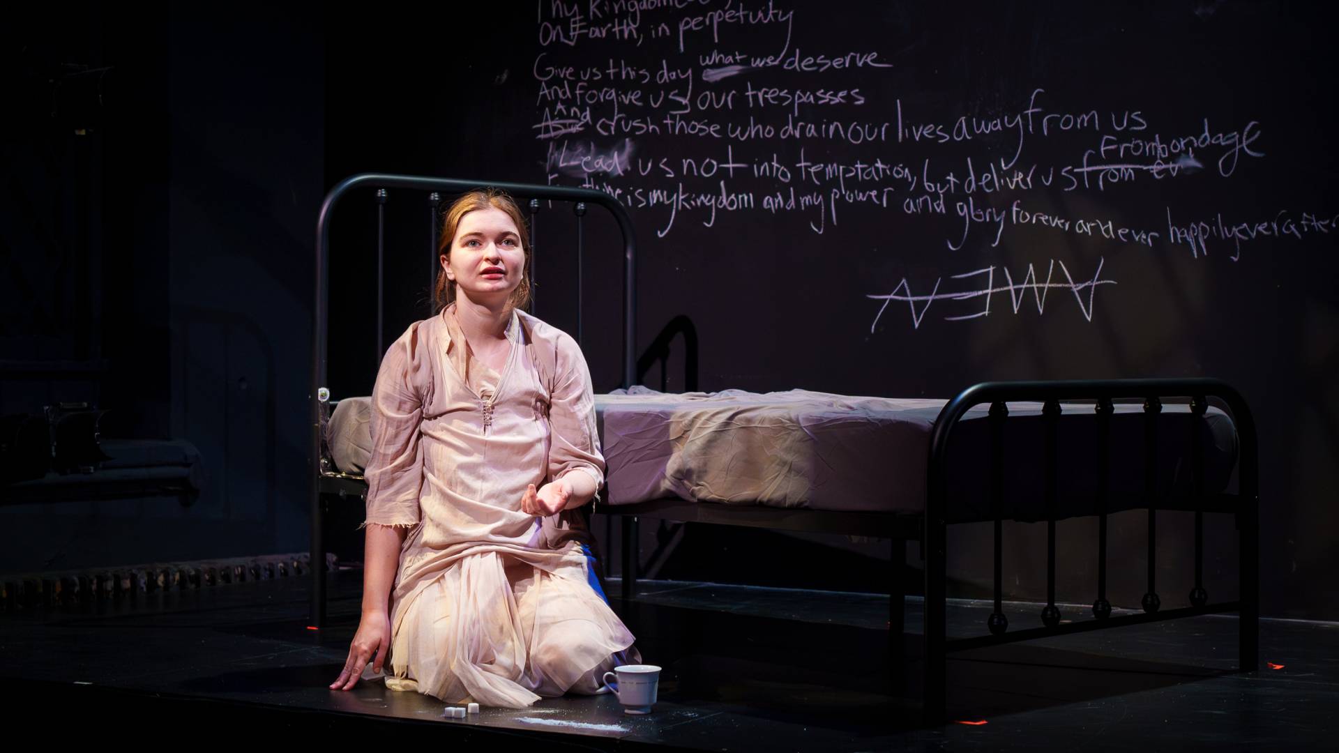 An actress recites a speech while sitting next to a wrought iron bed and the Lord's prayer scrawled in chalk behind her