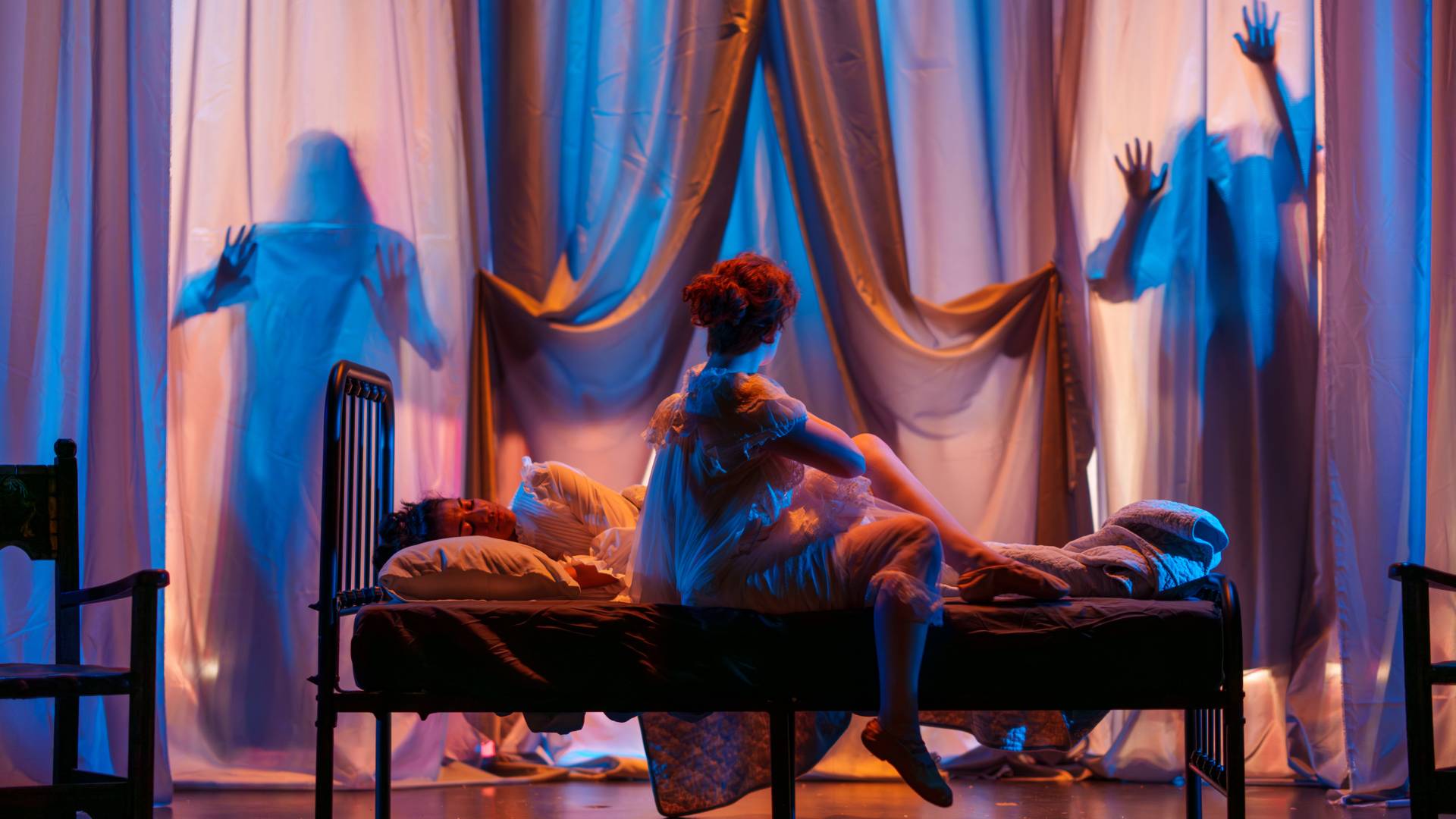 A girl sitting on a bed, looking at two silhouettes behind her.