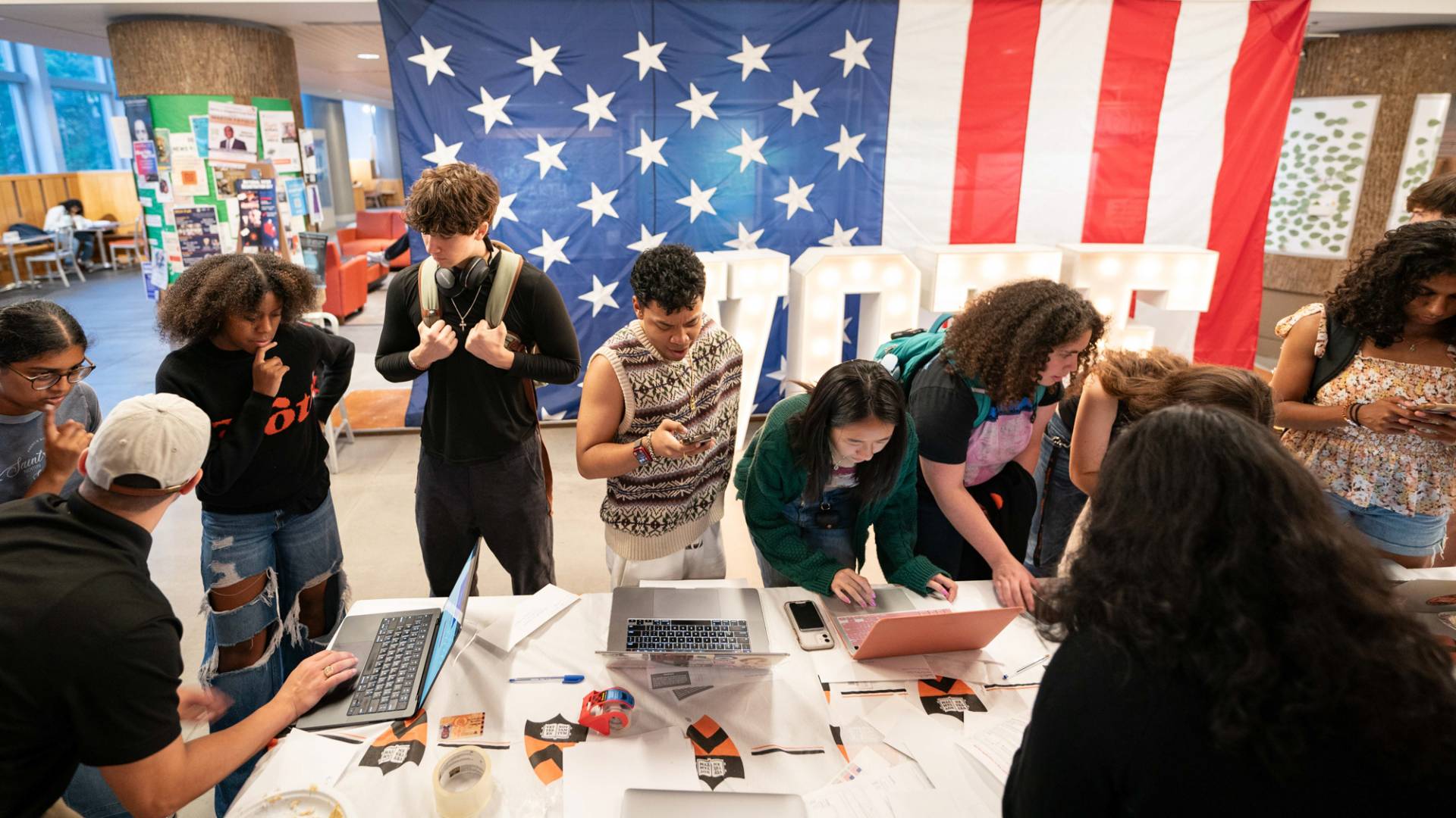 A group or people gathered around a table registering to vote, US flag in the background.