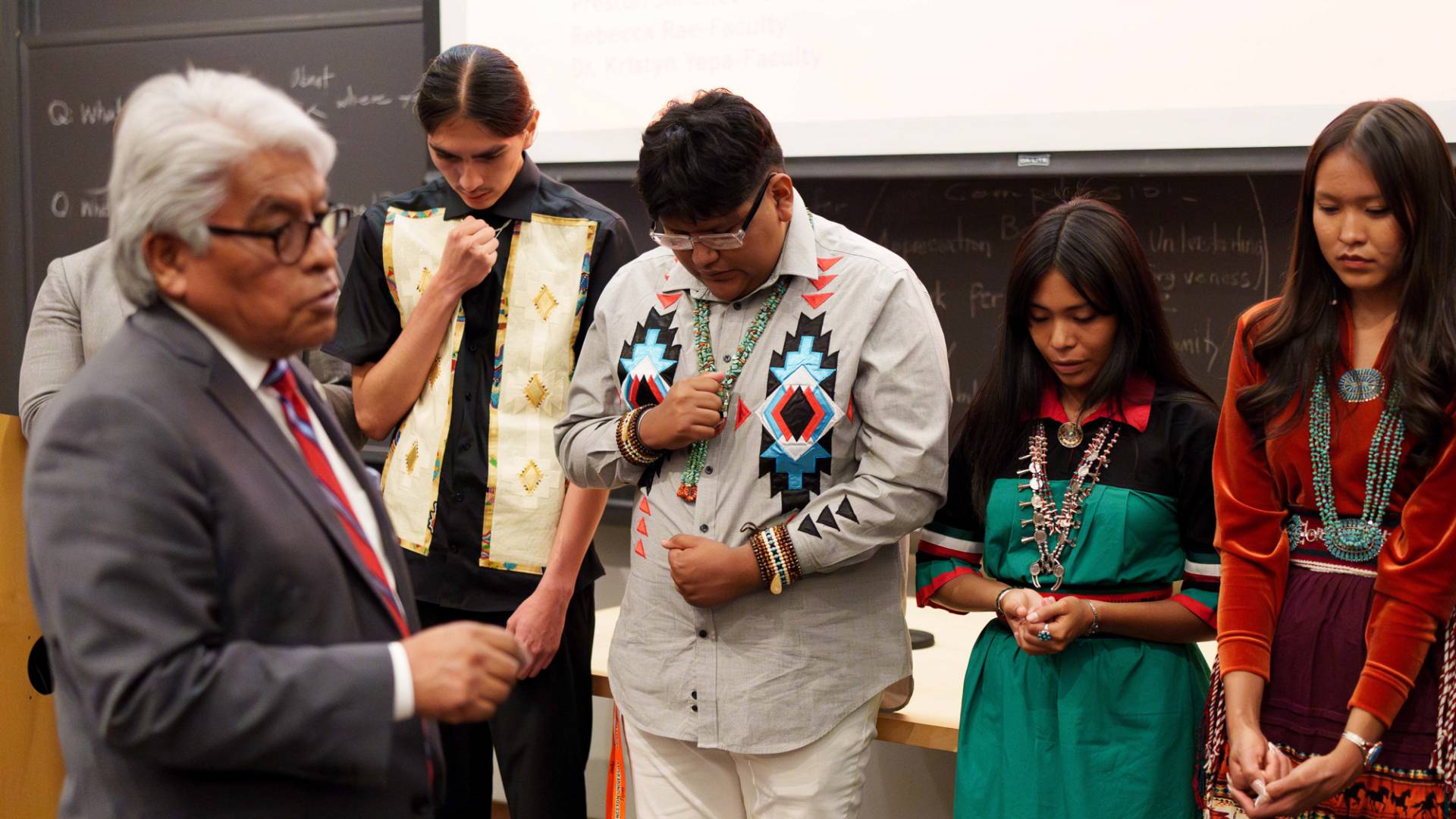 Native American students bowing, eyes closed standing shoulder to shoulder.