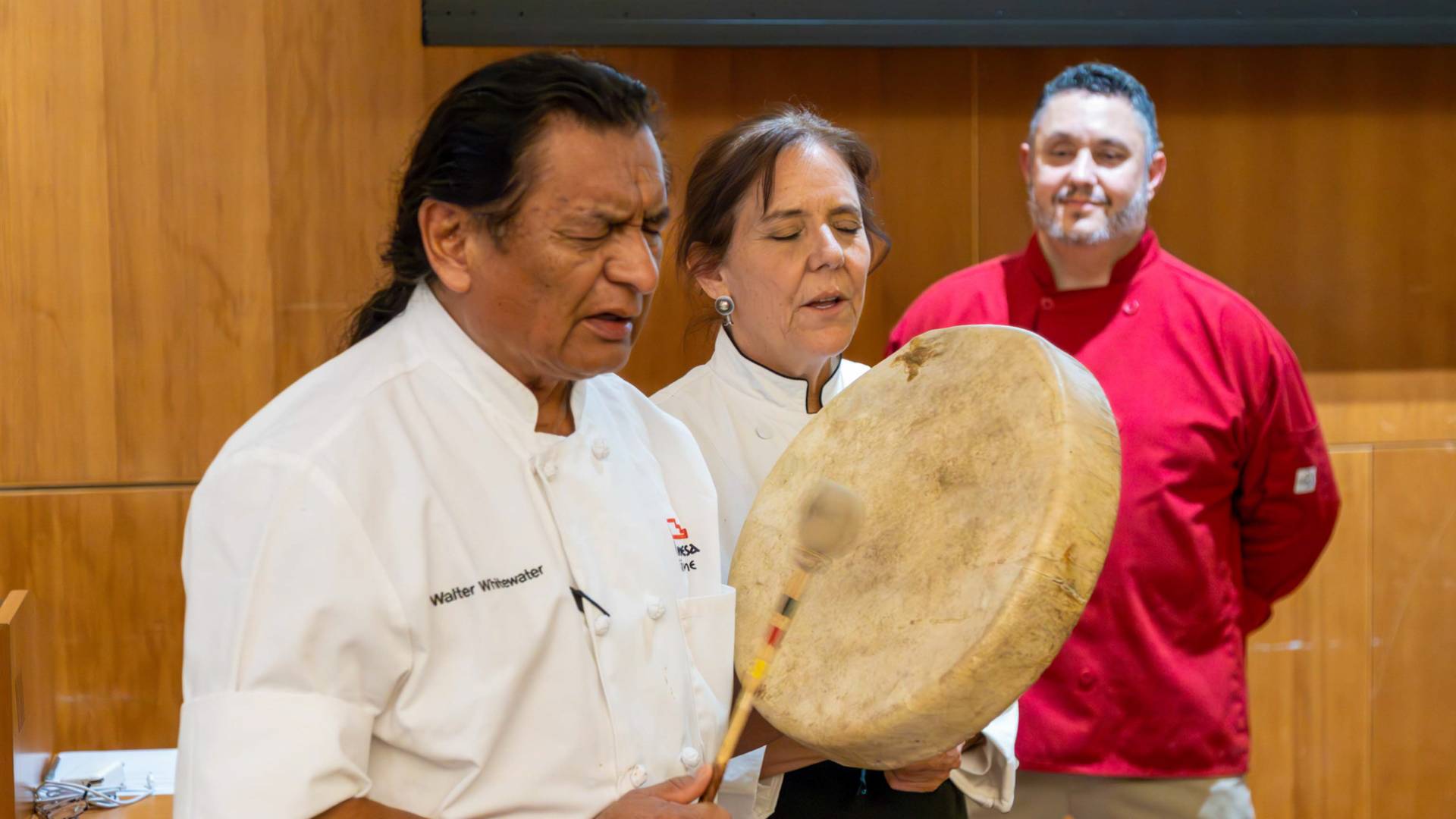 Native American dining staff member plays a drum with his eyes closed.