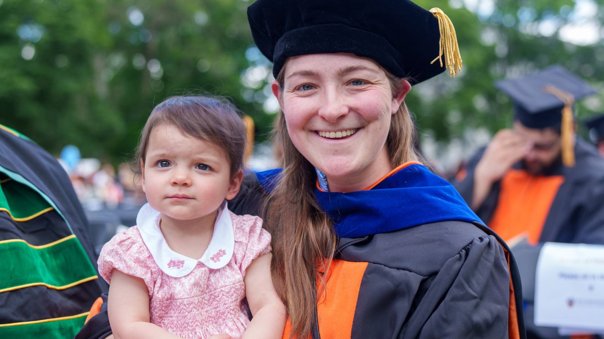 A Ph.D. graduate holds her daughter, in a pink dress.
