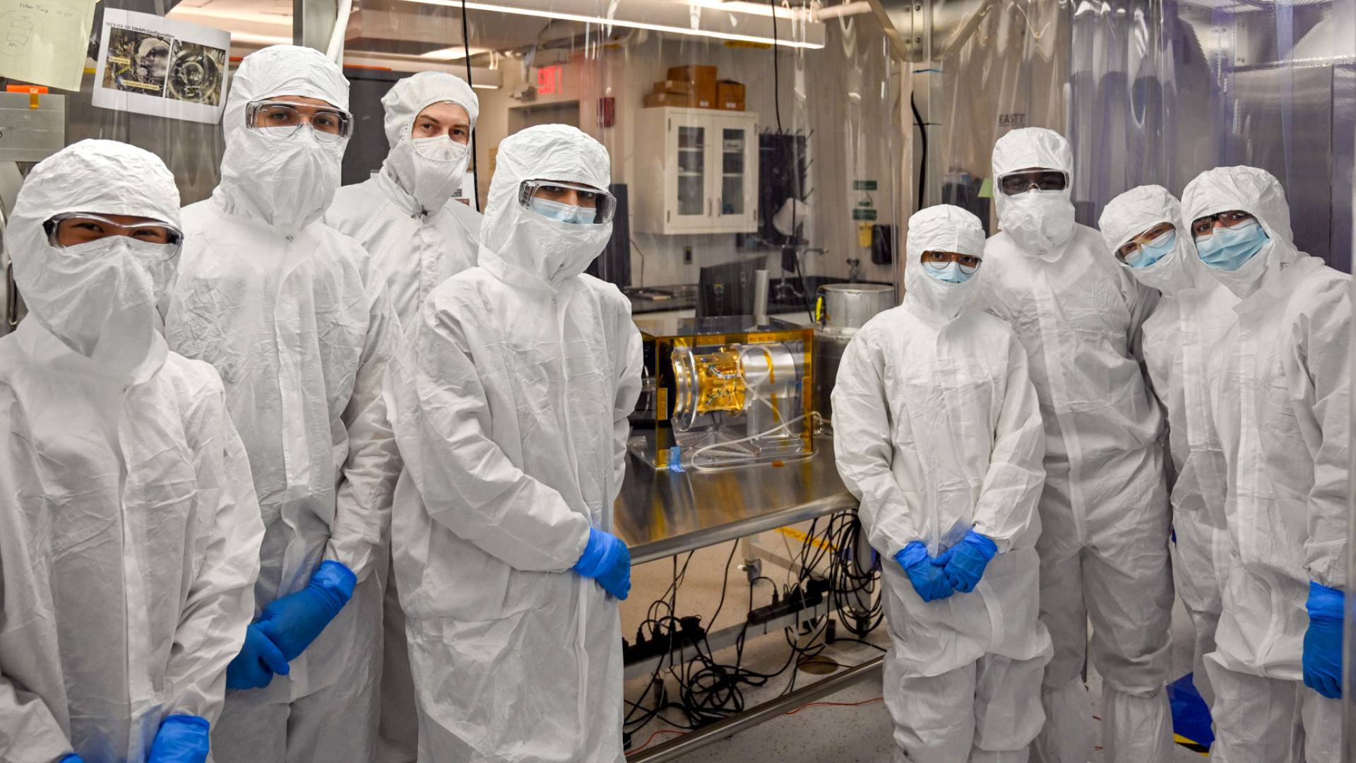 Eight people stand around the golden SWAPI instrument dressed in cleanroom gear.