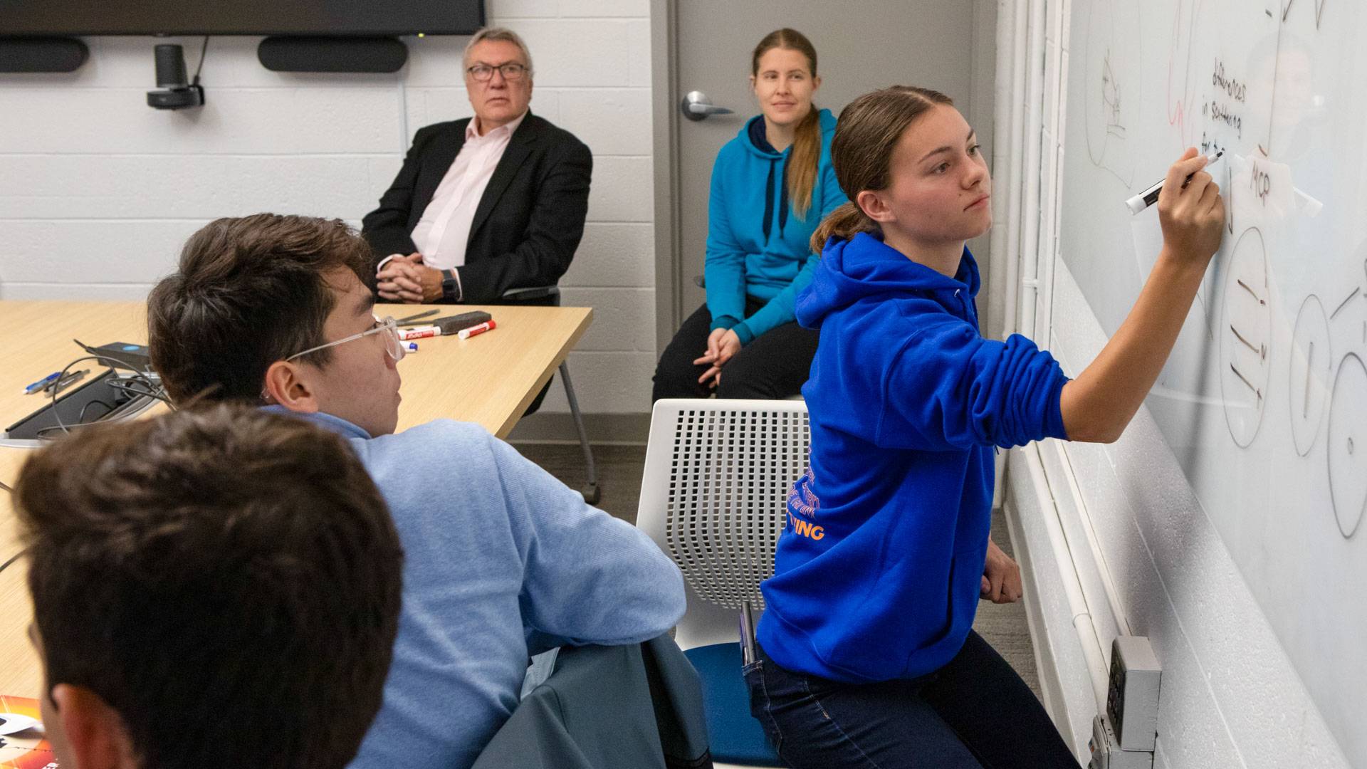 A student writes on a whiteboard as teachers and classmates look on.