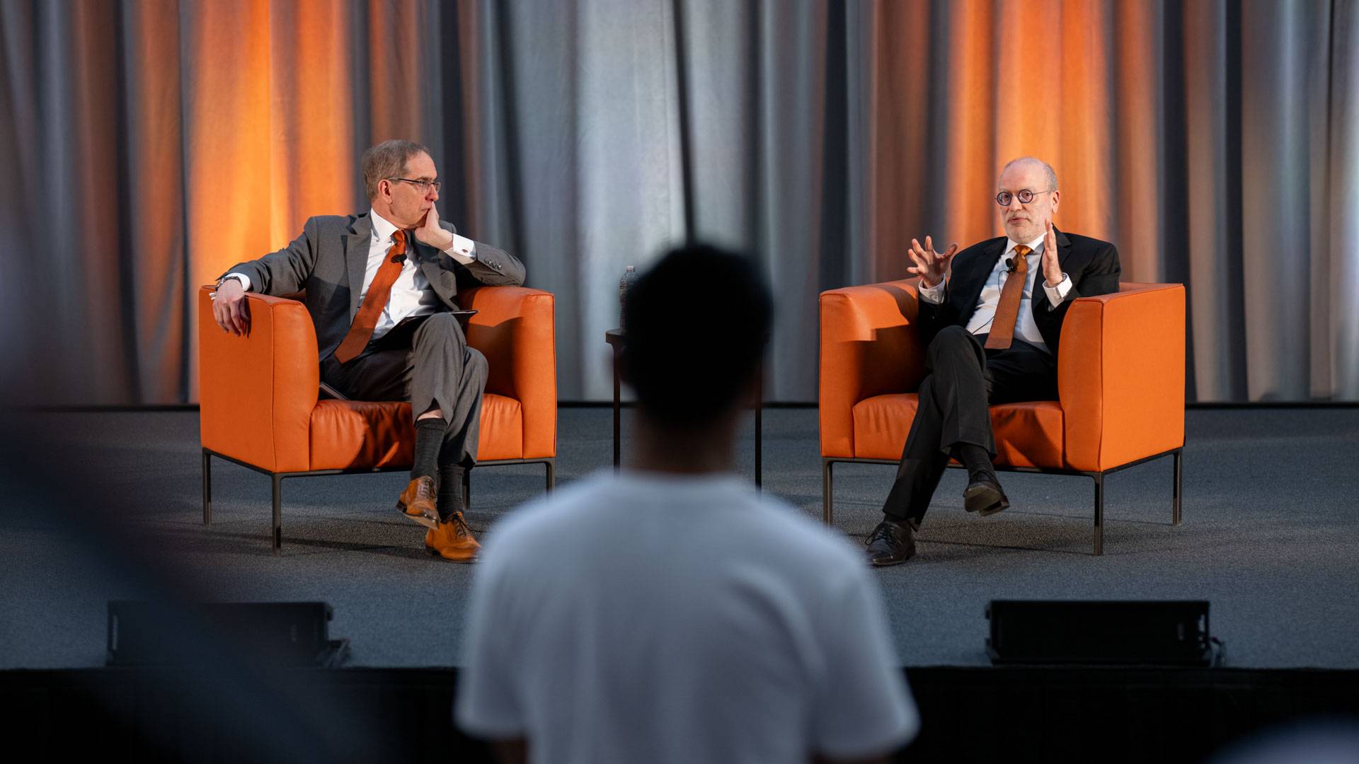 Two men in suits and orange ties sit in chairs on a stage in front of a group of students.