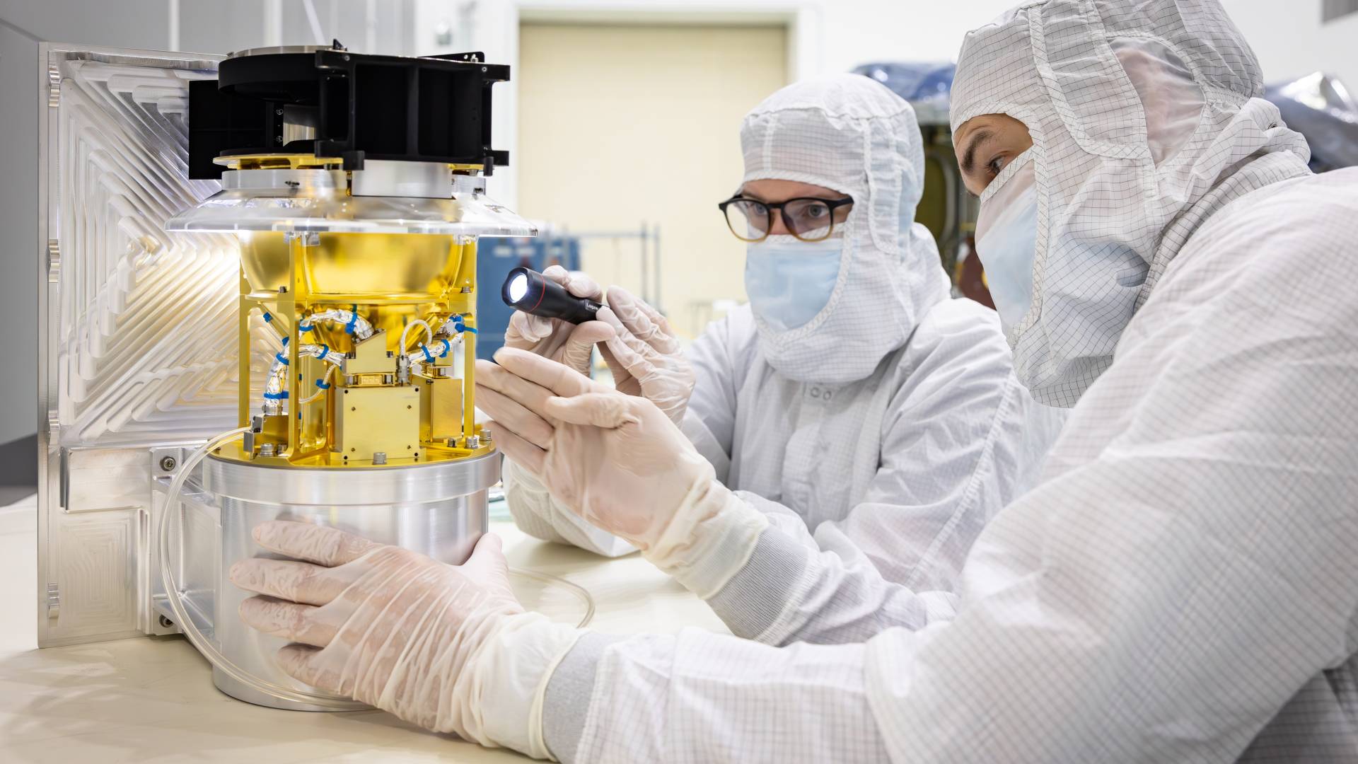 Scientists in cleanroom gear look at a golden instrument