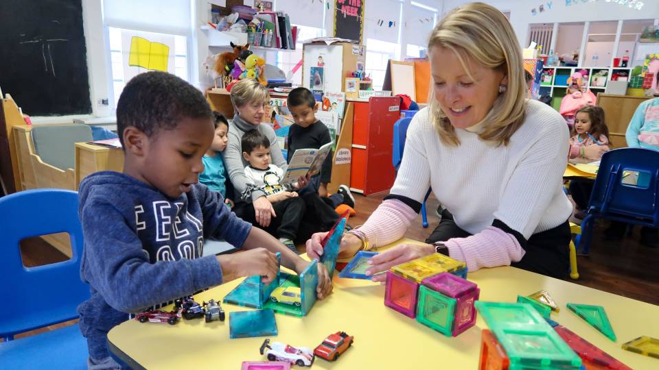Debby Foster sitting at table with Aiden playing with magnetic tiles. In background other four year olds being read to by Jennifer Caputo