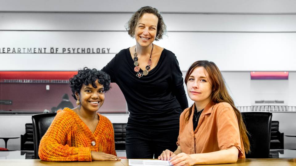 Student Kajal Schiller and postdoc Rachel Bedder sit at a table in the Department of Psychology with Professor Yael Niv standing between them.