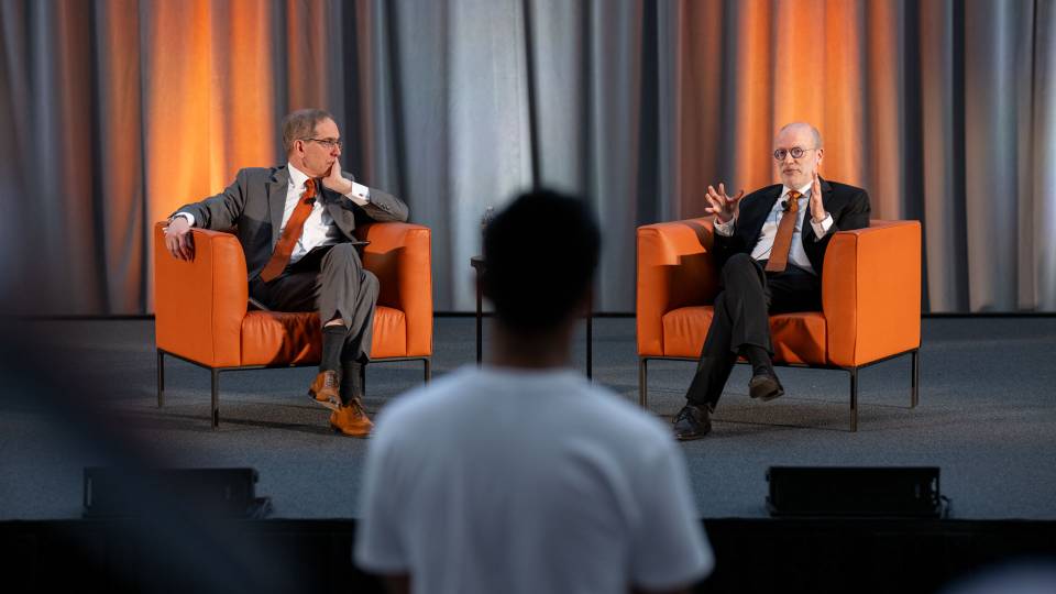 Two men in suits and orange ties sit in chairs on a stage in front of a group of students.