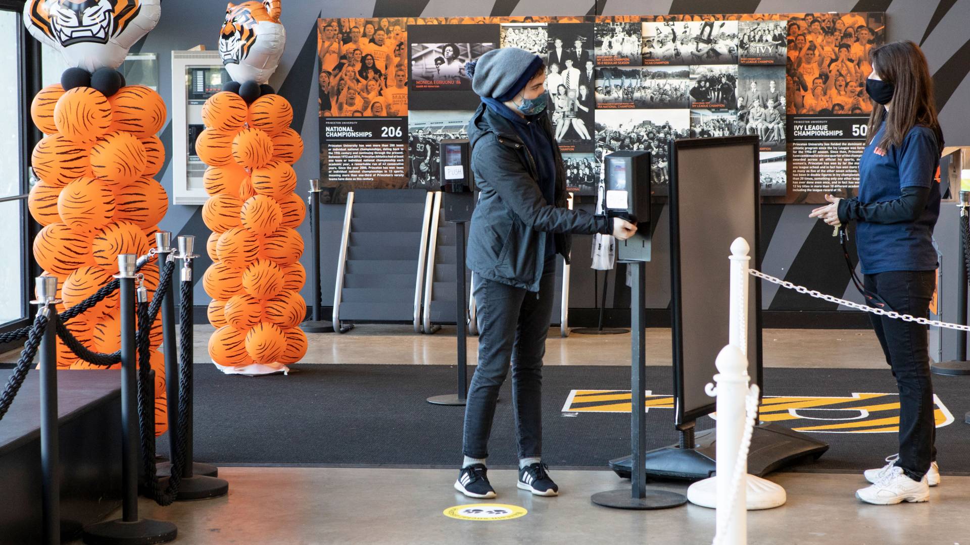 A student pumps hand sanitizer from a station while a staff member greets her
