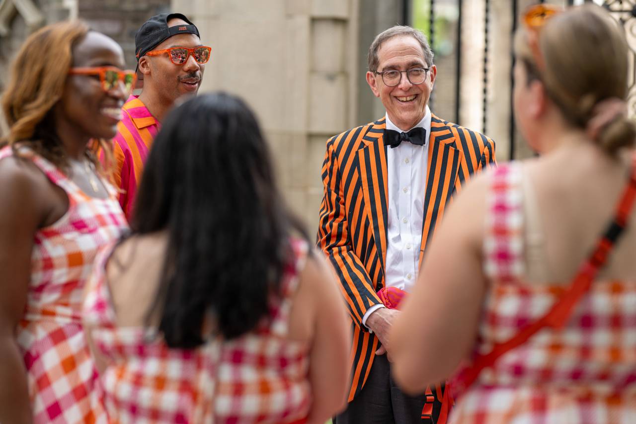 President Eisgruber wearing an orange and black jacket talking to a group of people.f