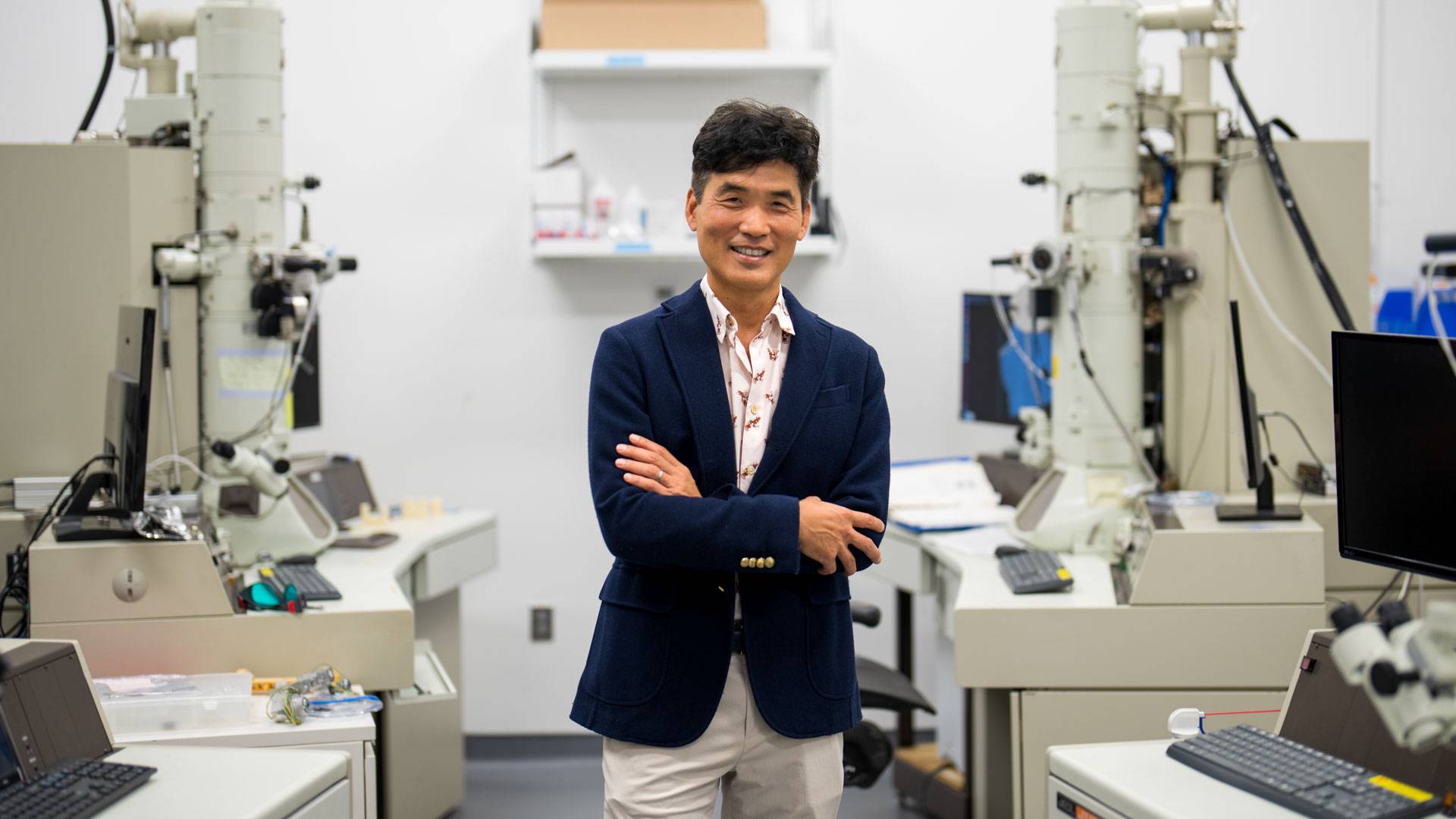 A handsome Korean American man leans against a large and complex machine.