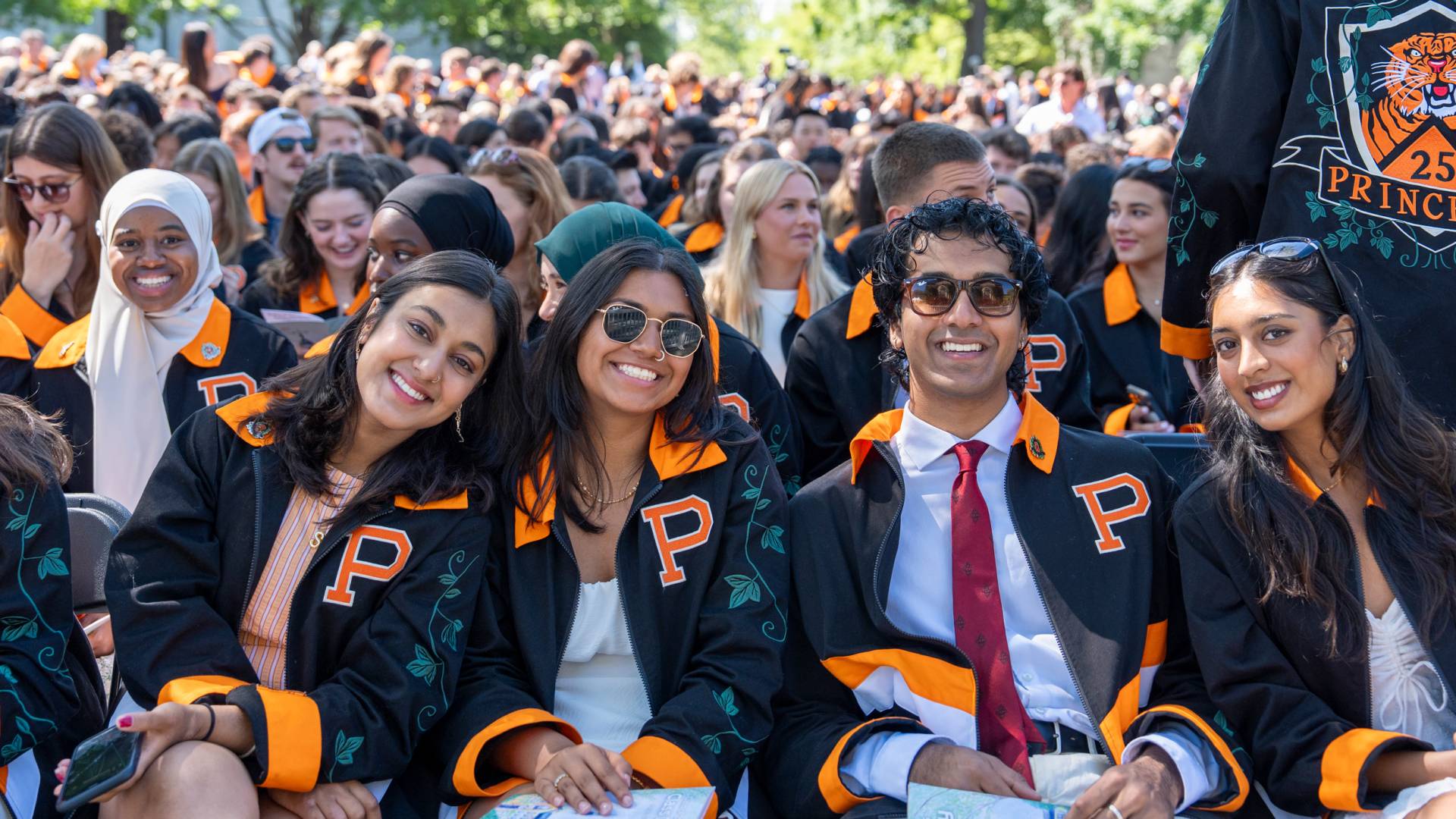 A smiling group of four Princeton seniors seated in front of the large Class Day audience.