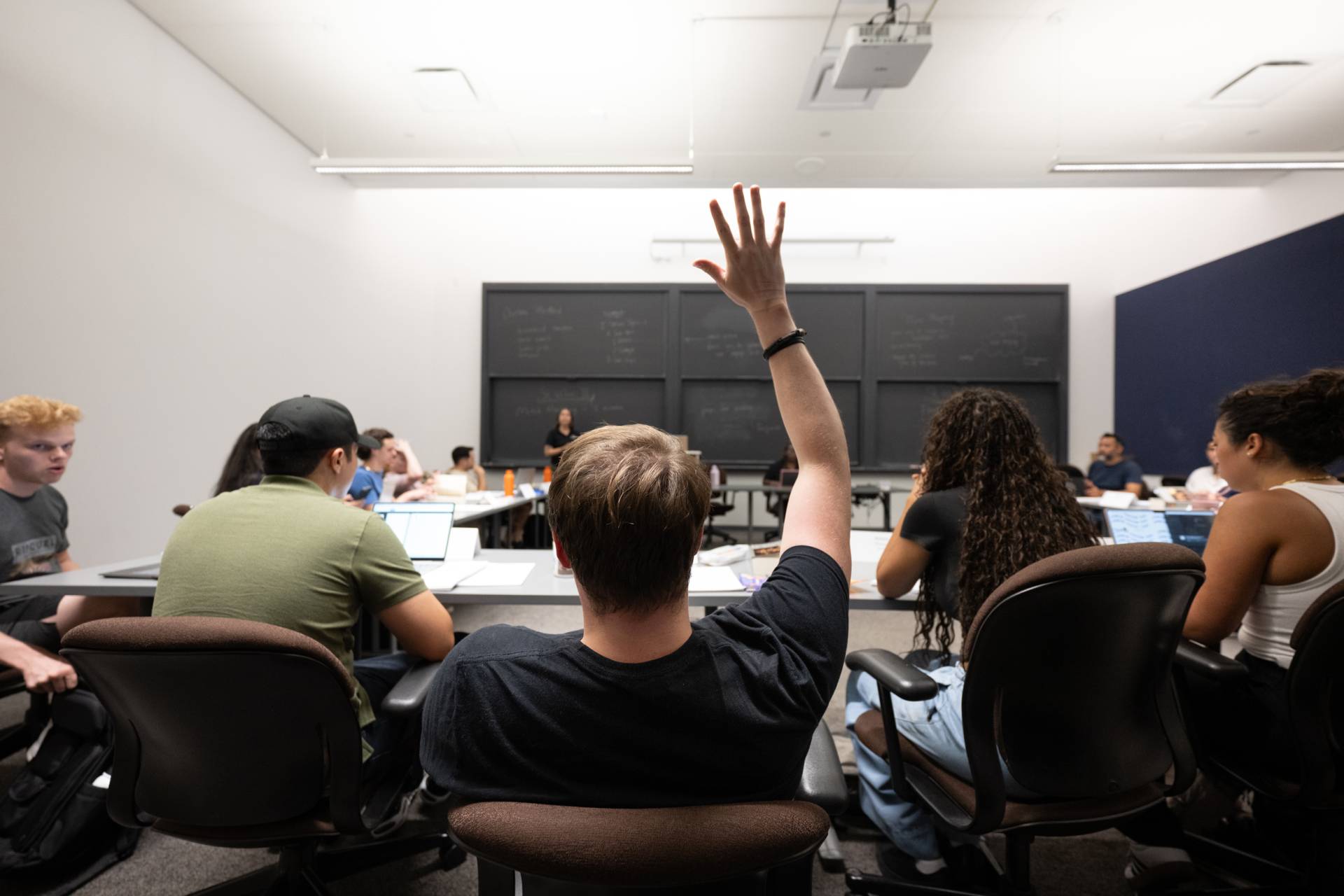 A student seated in the middle of a full classroom is pictured from the back raising their hand to ask a question.