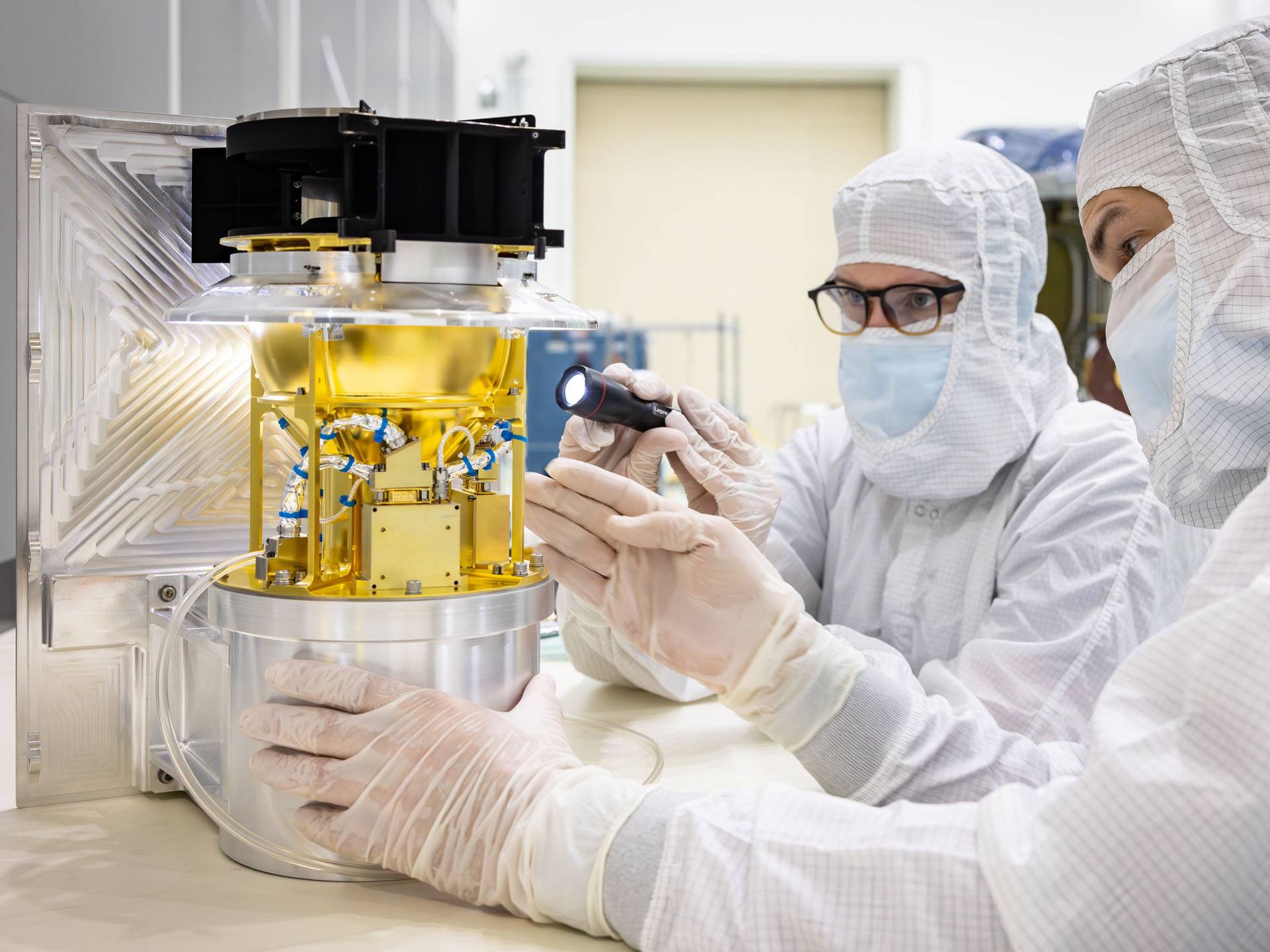 People in white coveralls perform delicate work on a golden piece of space craft instrumentation.