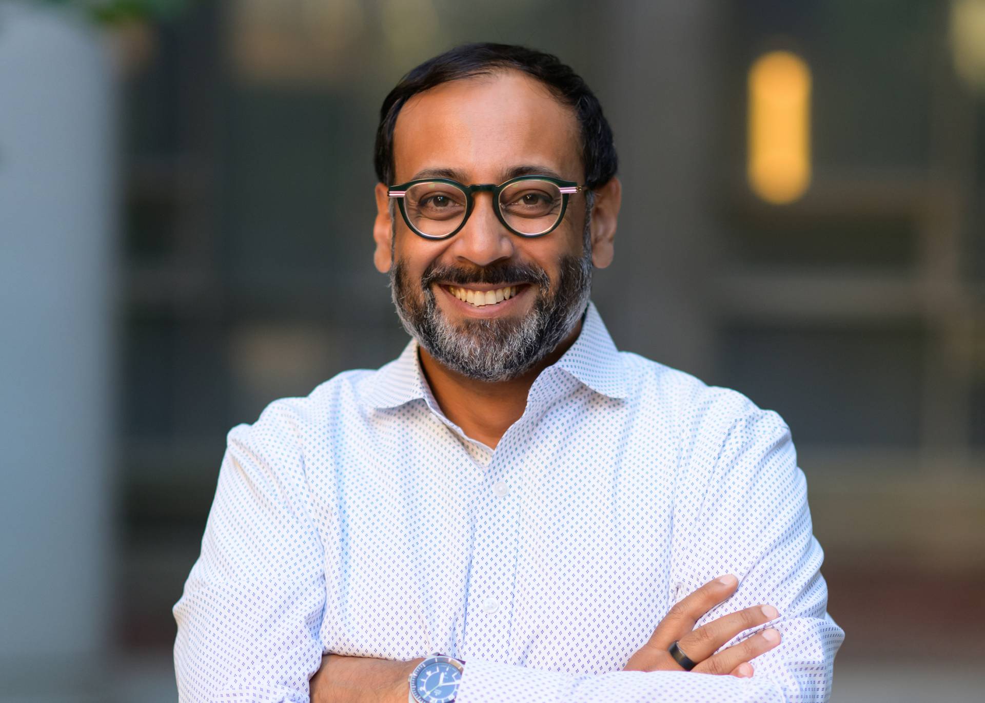 Nabarun Dasgupta smiles in a white dress shirt and glasses with short dark hair and beard smiling with his arms folded.