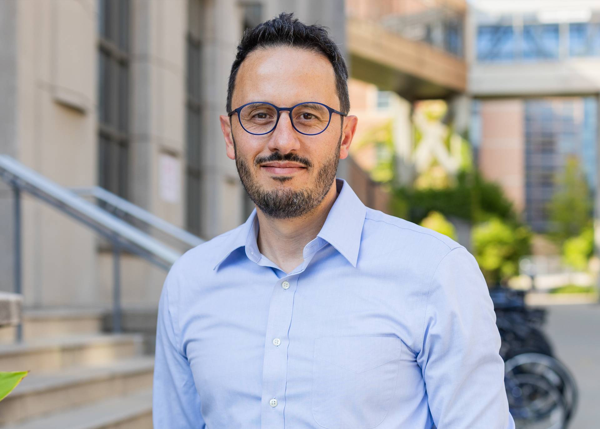 Man with light blue collared shirt, round frame glasses with dark short hair and a short mustache and beard smiling