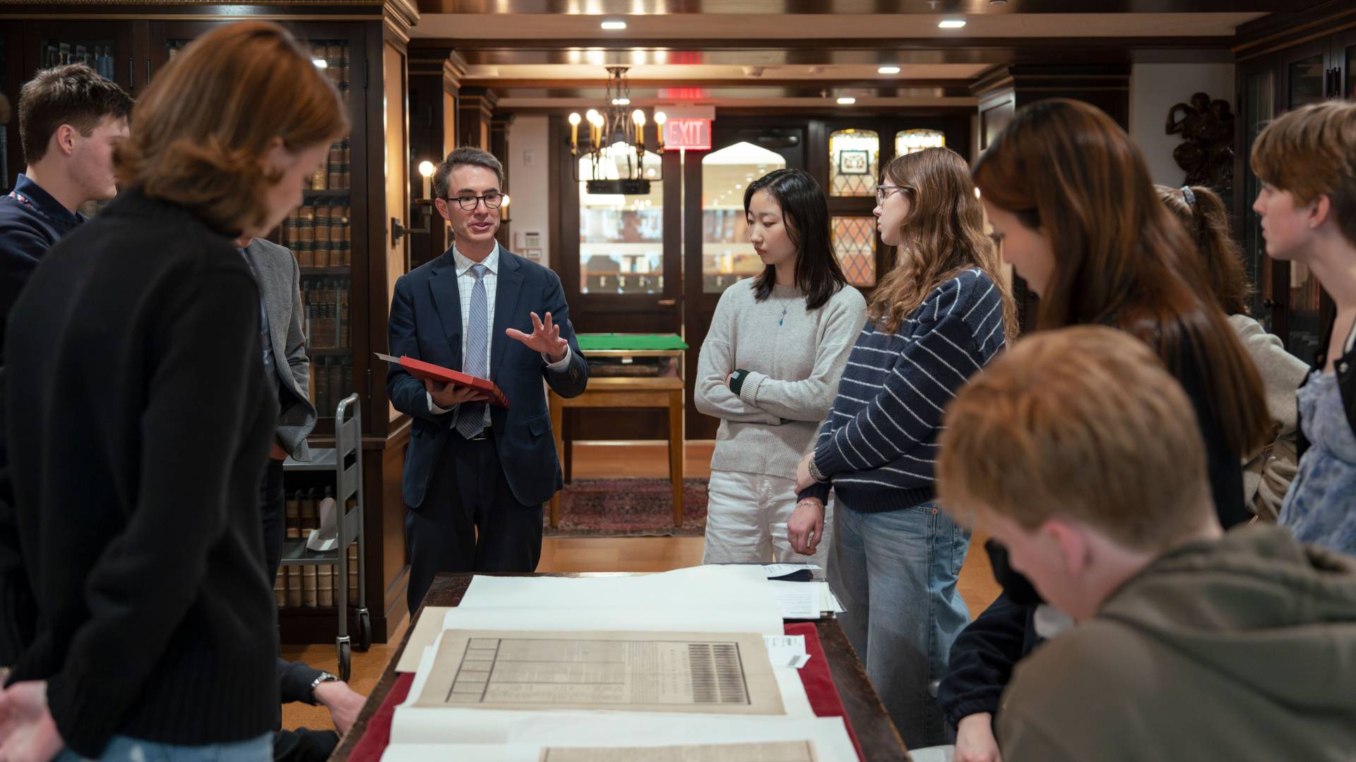 In a grand library, students listen intently to an instructor as he presents on formats of communication in the revolutionary era