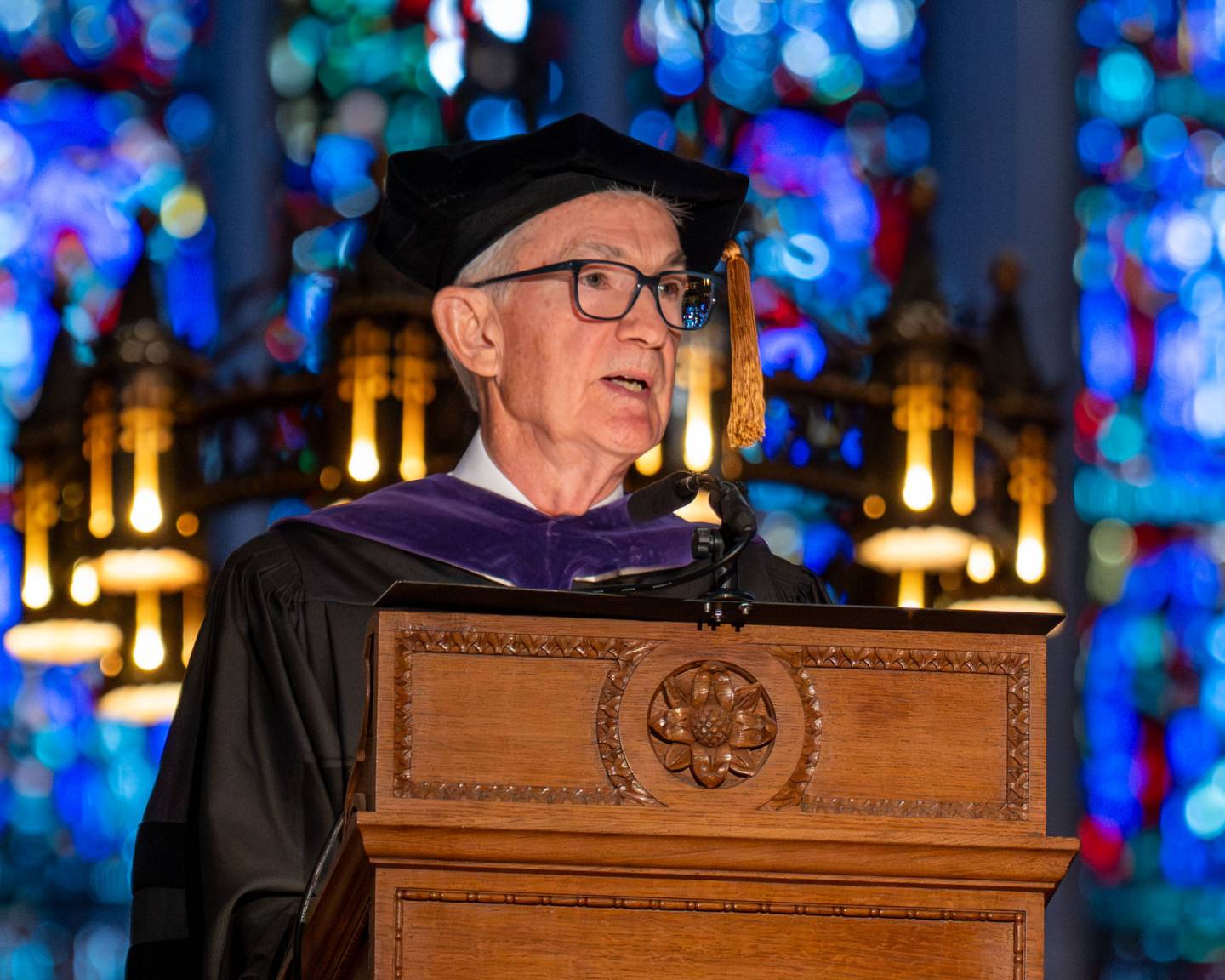 A man in graduation regalia speaks at a podium in front of stained glass.